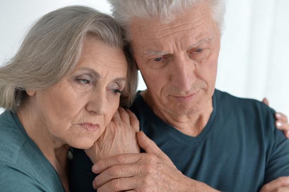 Ein älteres Paar steht eng umschlungen, beide blicken nachdenklich und wirken emotional berührt.